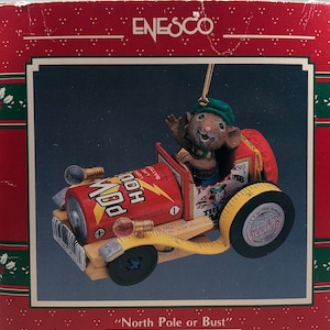 May include: A red, white, and blue Christmas ornament featuring a mouse driving a toy car made from a can of popcorn. The car has a tape measure for a tire and a "Golly Gee Wiz" logo on the side. The ornament reads "North Pole or Bust".