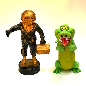 Può includere: Una statuetta di un palombaro con casco dorato e tuta nera che tiene in mano uno scrigno dorato, su una base circolare nera, accanto a una statuetta di coccodrillo verde con lingua rossa e occhi grandi.