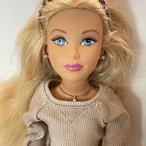 Könnte beinhalten: Puppe mit blonden Haaren, blauen Augen und pinkfarbenem Lippenstift. Die Puppe trägt ein beiges geripptes Langarmshirt und eine goldene Halskette. Ein braunes Stirnband befindet sich auf dem Kopf der Puppe. Die Puppe ist ein Sammlerstück.