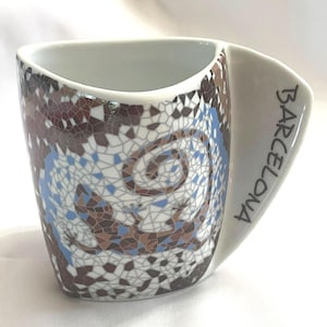 May include: White ceramic mug with a mosaic design in brown, blue, and white. The handle is white with the word "BARCELONA" printed in black. The mug has a unique, angular shape.