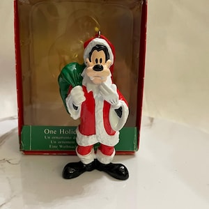 May include: A red and white Santa Claus ornament featuring Goofy holding a green bag. The ornament is in its original box with the text "One Holiday" on the front.