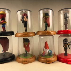 Puede incluir: Ocho c&uacute;pulas de pl&aacute;stico transparente con bases negras, rojas y marrones, cada una con una peque&ntilde;a figura de pl&aacute;stico. Las figuras incluyen un gnomo, una abeja, una ni&ntilde;a con cabello rojo, una ni&ntilde;a con cabello marr&oacute;n, un ni&ntilde;o con un sombrero rojo, un ni&ntilde;o con un sombrero amarillo, un robot y una ni&ntilde;a con un sombrero rojo.