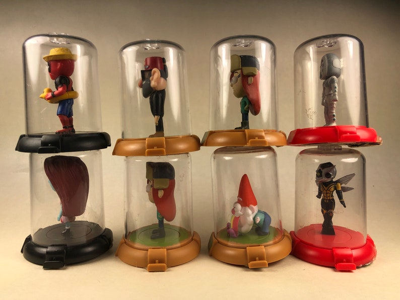 Puede incluir: Ocho c&uacute;pulas de pl&aacute;stico transparente con bases negras, rojas y marrones, cada una con una peque&ntilde;a figura de pl&aacute;stico. Las figuras incluyen un gnomo, una abeja, una ni&ntilde;a con cabello rojo, una ni&ntilde;a con cabello marr&oacute;n, un ni&ntilde;o con un sombrero rojo, un ni&ntilde;o con un sombrero amarillo, un robot y una ni&ntilde;a con un sombrero rojo.