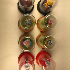 Puede incluir: Ocho contenedores de pl&aacute;stico transparente con bases rojas, amarillas y verdes. Cada contenedor tiene una peque&ntilde;a figura de pl&aacute;stico en su interior. Las figuras son de varios personajes, incluyendo un conejo, un gato, un perro y un p&aacute;jaro. Los contenedores est&aacute;n etiquetados con la palabra "Glitz" en el lateral.