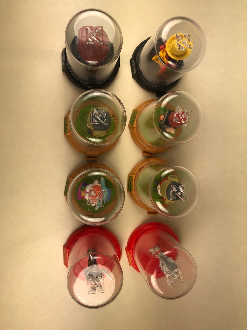 Puede incluir: Ocho contenedores de pl&aacute;stico transparente con bases rojas, amarillas y verdes. Cada contenedor tiene una peque&ntilde;a figura de pl&aacute;stico en su interior. Las figuras son de varios personajes, incluyendo un conejo, un gato, un perro y un p&aacute;jaro. Los contenedores est&aacute;n etiquetados con la palabra "Glitz" en el lateral.