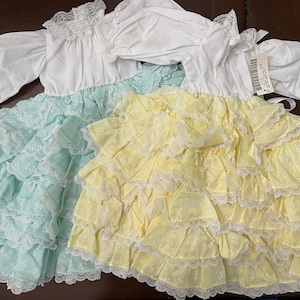 Puede incluir: Dos vestidos blancos con volantes y ribete de encaje. Un vestido es azul claro y el otro es amarillo.