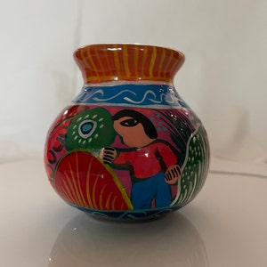 May include: A small, round, hand-painted ceramic vase with a colorful design. The vase features a person in a red shirt and blue pants, surrounded by green and red foliage. The vase has a blue and white rim.