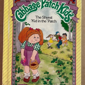 May include: A book cover featuring a Cabbage Patch Kid doll with red hair and a purple dress. The doll is holding a basket of strawberries. The book title is "The Shyest 'Kid in the 'Patch" and is a Parker Brothers Story Book.