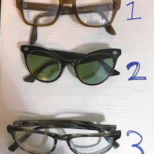 Puede incluir: Se muestran tres pares de gafas. El par superior tiene monturas cuadradas marrones. El par del medio presenta monturas negras de ojo de gato con lentes verdes. El par inferior tiene monturas grises y transparentes con forma rectangular.