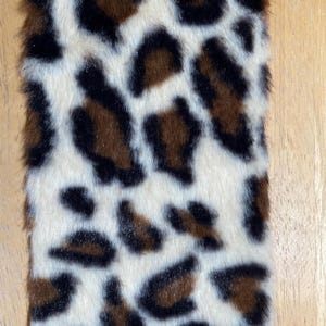 May include: Close-up of a faux fur textile with a leopard print pattern. The design features irregular black and brown spots on a cream-colored background. The fur has a soft, textured appearance, suggesting a plush material.
