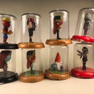 Puede incluir: Ocho peque&ntilde;as figuritas se exhiben en c&uacute;pulas de pl&aacute;stico transparente sobre una superficie blanca. Las figuritas son de varios colores y representan personajes de pel&iacute;culas y programas de televisi&oacute;n populares. Las c&uacute;pulas est&aacute;n dispuestas en fila, con las figuritas mirando hacia adelante.