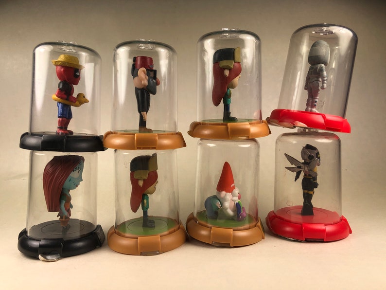 Puede incluir: Ocho peque&ntilde;as figuritas se exhiben en c&uacute;pulas de pl&aacute;stico transparente sobre una superficie blanca. Las figuritas son de varios colores y representan personajes de pel&iacute;culas y programas de televisi&oacute;n populares. Las c&uacute;pulas est&aacute;n dispuestas en fila, con las figuritas mirando hacia adelante.