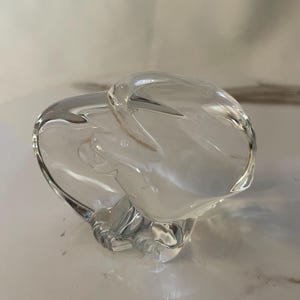 May include: A clear glass sculpture of an abstract shape. The sculpture is smooth and has a rounded, organic form.