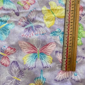 May include: Purple fabric with a repeating pattern of colourful butterflies. The fabric has a glittery texture. A wooden ruler with inch markings is partially visible in the bottom right corner.