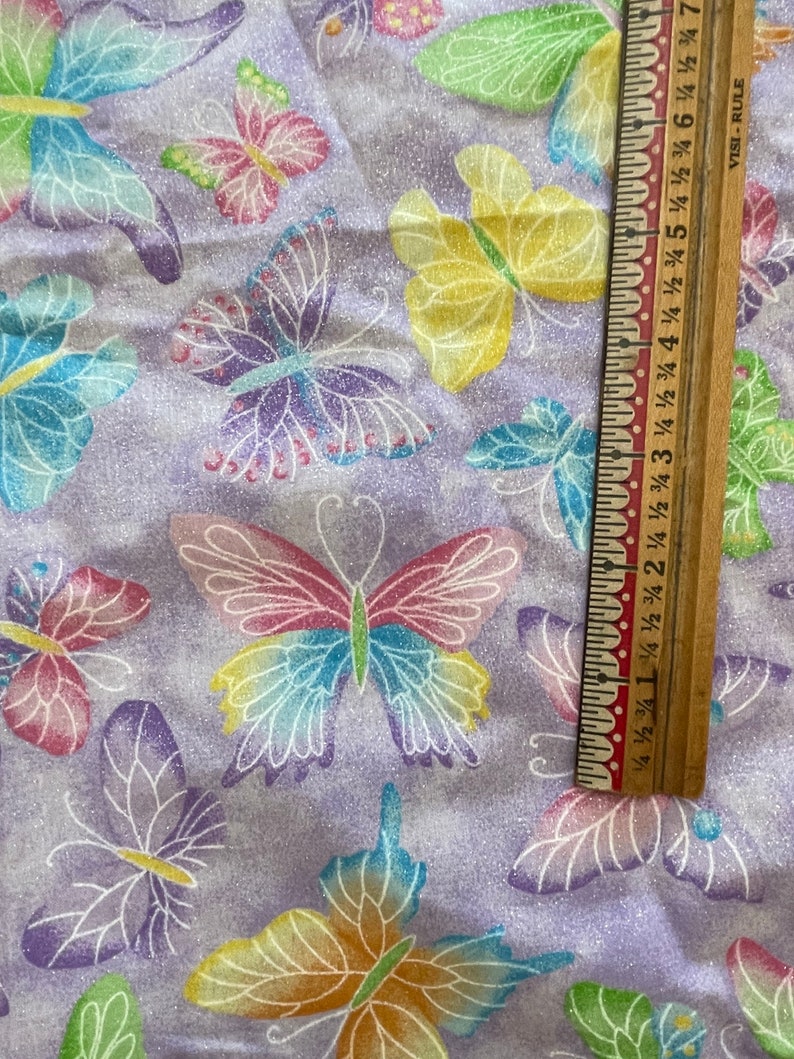 May include: Purple fabric with a repeating pattern of colourful butterflies. The fabric has a glittery texture. A wooden ruler with inch markings is partially visible in the bottom right corner.
