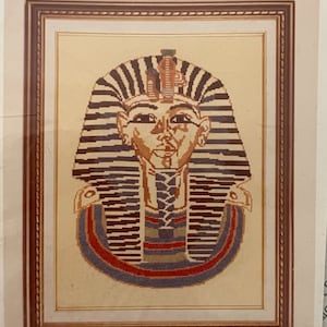 May include: A framed cross-stitch of a pharaoh's mask with a striped headdress, red and blue accents, and a beige background.