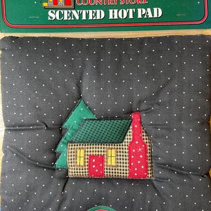 May include: Black, polka-dotted, quilted hot pad with a fabric applique of a house and tree. The house has a red chimney and red and yellow accents. The packaging reads "Sarah Lynn's Country Store Scented Hot Pad."