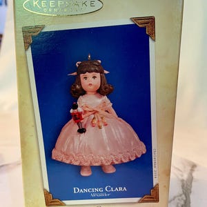May include: Hallmark Keepsake Ornament box featuring a doll named Dancing Clara. The doll is wearing a pink dress and holding a toy soldier. The box has gold accents and the text "Collector's Series".