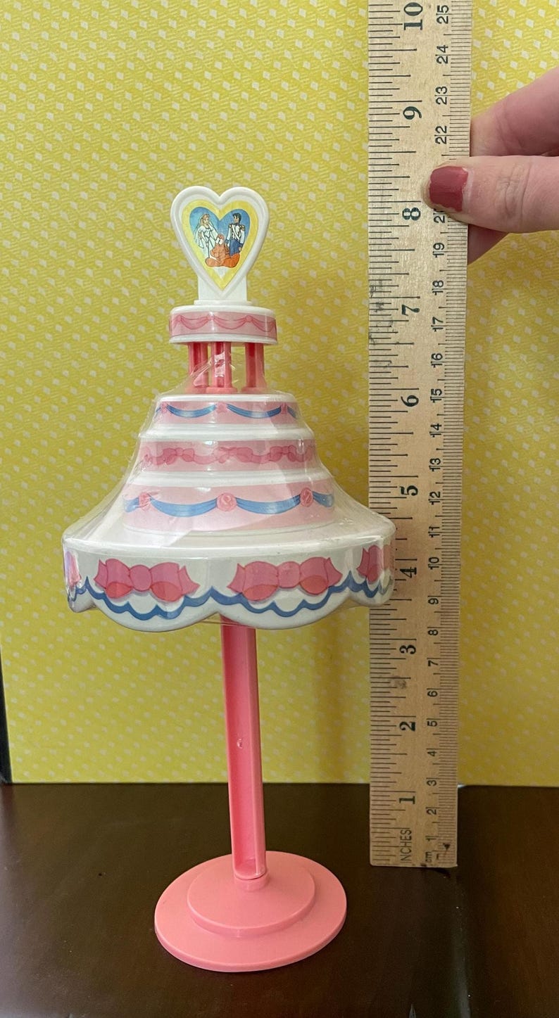 以下が含まれることがあります： ピンク色のプラスチック製のケーキ型おもちゃ。ハート型のトッパーにはカップルのイラストが描かれています。ケーキにはピンクと青の装飾が施されています。おもちゃの高さは約23cmです。