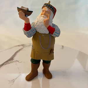 May include: A Santa Claus figurine wearing a brown and green outfit with a yellow apron and a red and white striped shirt. He is holding a small wooden boat in his left hand and has a thoughtful expression on his face.