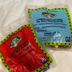 May include: Two sealed McDonald's Happy Meal Cabbage Patch Kids toys. One package is red with green trim and the other is blue with green trim. Both packages have text that reads "Congratulations On Your New Arrival!"