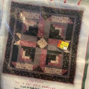May include: A Learn to Quilt Log Cabin wall hanging kit in a plastic package. The quilt design features a square pattern with dark brown, red, and cream floral fabrics. The packaging text includes 76.2 cm by 76.2 cm wall hanging.