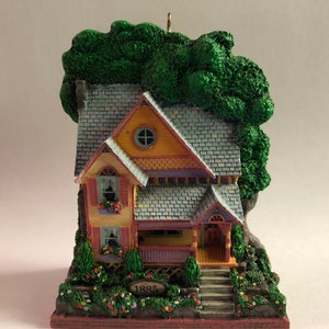 May include: A miniature house ornament with a yellow, blue, and red exterior, a green tree, and a porch with steps. The house has a small window and the date "1885" is inscribed on a plaque.
