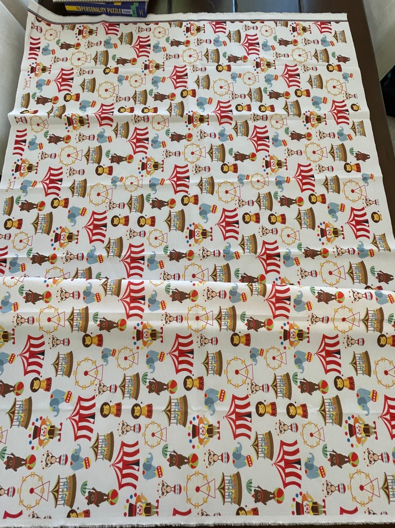 May include: White fabric with a repeating pattern of a circus theme. The pattern includes red and white striped tents, elephants, monkeys, clowns, Ferris wheels, and other circus elements.