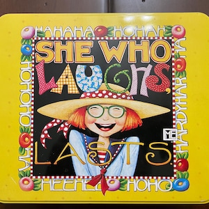 Puede incluir: Una lata rectangular de metal amarillo con una ilustración central de una persona con cabello rojo, gafas verdes y sombrero de paja. El texto dice SHE WHO LAUGHS LASTS, rodeado por un borde de frutas coloridas y la palabra HAHAHA repetida.