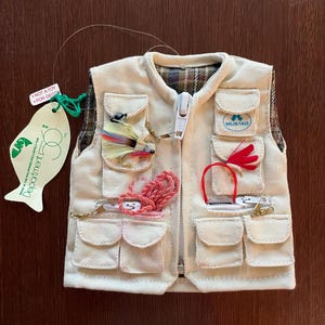 May include: A miniature beige fishing vest with multiple pockets and a zipper. The vest is adorned with fishing lures and a tag that reads "NOT A TOY FOR DECOR". The vest has the brand name "MUSTAD" on one of the pockets.