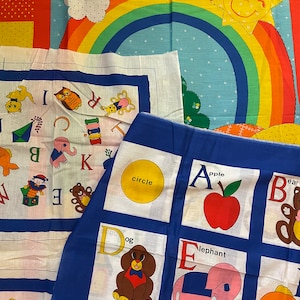 May include: A blue fabric alphabet chart with colorful illustrations of objects for each letter. The chart includes a circle, apple, dog, elephant, ice cream, and a fish. The letters are in a white box with a blue border.