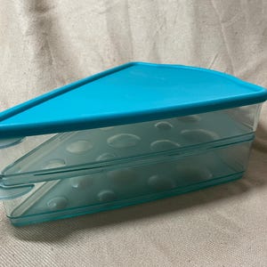 May include: A clear, triangular food storage container with a turquoise lid. The container has two stacked sections with a series of small, rounded indentations. The lid and base have a matching turquoise color. The container is designed to store food.