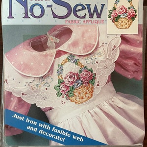 May include: A pink and white dress with a white lace collar and a floral applique of a basket of roses. The package includes instructions for using fusible web to iron on the applique. The text on the package reads "Daisy Kingdom Mini No-Sew Fabric Applique #C113-1680-007 Rose Bouquet".
