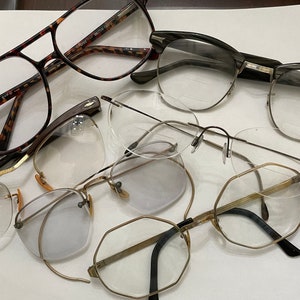 May include: A collection of five pairs of eyeglasses with various frame styles and colors. The eyeglasses are arranged on a white surface.