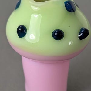 May include: A mushroom-shaped glass object with a light green top and a pink base. The top has dark blue spots and a blue rim. The object is likely a decorative piece or a functional item.
