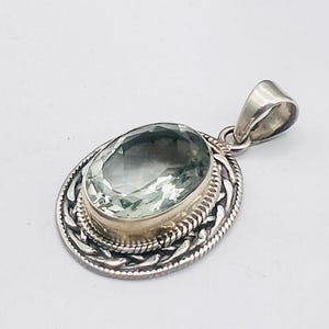 May include: An oval-shaped pendant featuring a faceted, light green gemstone set in a silver bezel. The setting is surrounded by a decorative, twisted rope-style border and a textured metal frame, complete with a polished silver bail at the top.