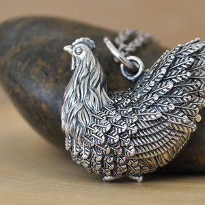 May include: A silver pendant necklace featuring a detailed, three-dimensional design of a hen with its wings spread. The pendant is made of metal and has a textured, feathered look.
