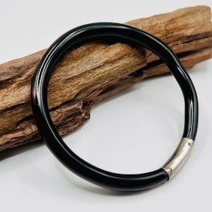 May include: A glossy black bangle bracelet with a silver clasp. The bracelet is circular and appears to be made of a solid material. The bracelet is resting on a piece of driftwood.