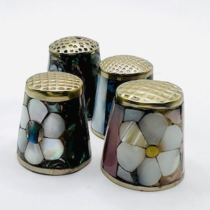 May include: A set of four sewing thimbles featuring metallic tops with a textured grid pattern. Each thimble is decorated with inlaid iridescent shell pieces, including white floral motifs on dark and light backgrounds.