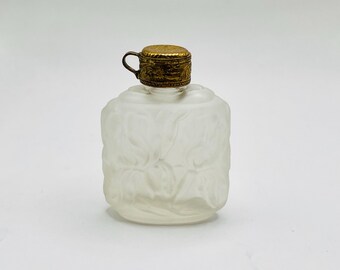 Può includere: Un flacone di profumo in vetro smerigliato con un motivo floreale decorativo. Il flacone ha un coperchio in metallo color oro con una piccola maniglia. Il flacone è rettangolare con bordi arrotondati e una finitura smerigliata.