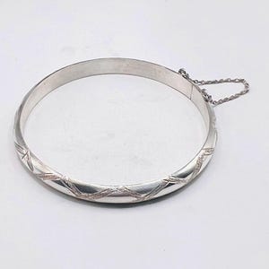May include: A polished silver-tone hinged bangle bracelet featuring a repeating etched crisscross pattern along the outer surface. The circular bracelet includes a secure clasp mechanism and a delicate attached safety chain for added security.