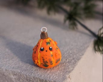 Puede incluir: Un pequeño adorno con forma de calabaza naranja con una cara alegre, ojos negros y una pequeña sonrisa. El adorno tiene detalles verdes y amarillos y un tallo marrón, perfecto para la decoración de otoño o Halloween.
