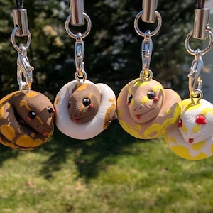 May include: Four small, colourful, handmade polymer clay snake keyrings. Each keyring features a different colour pattern, including brown and yellow, white and brown, tan and yellow, and yellow and white.