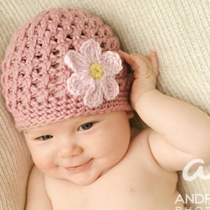 May include: A baby wearing a pink crocheted hat with a flower applique. The hat is a soft, light pink color, and the flower has a yellow center. The baby is lying on a cream-colored blanket.