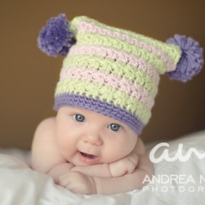 May include: A baby wearing a green, pink and purple striped crocheted hat with pom-poms. The baby is lying on a white surface and looking at the camera.