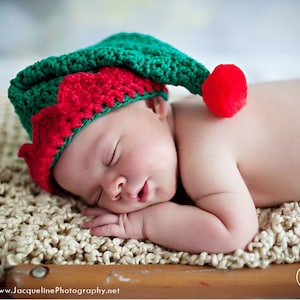 May include: A newborn baby wearing a green and red crocheted elf hat with a red pom-pom on top, sleeping on a beige crocheted blanket.