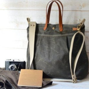 May include: A large, dark green canvas tote bag with brown leather handles and a long, adjustable strap. The bag is sitting on a wooden surface with a camera, a book, and a pair of pants.