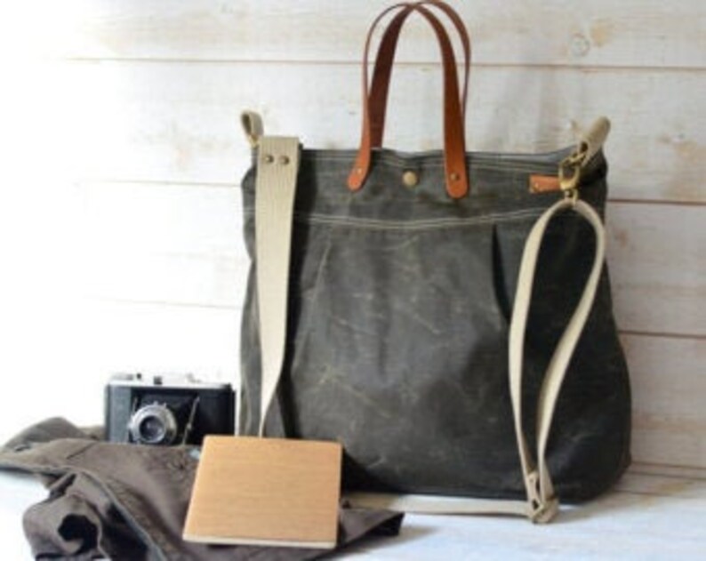 May include: A large, dark green canvas tote bag with brown leather handles and a long, adjustable strap. The bag is sitting on a wooden surface with a camera, a book, and a pair of pants.