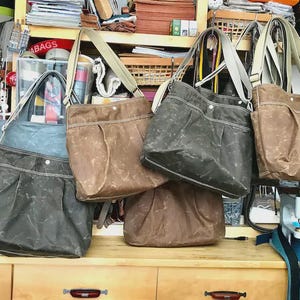May include: A collection of five shoulder bags in various shades of brown, gray, and blue, each featuring a wide shoulder strap, a pleated front pocket, and a single snap button closure, displayed on a wooden workbench in a workshop setting.