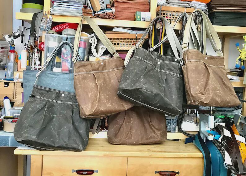 May include: A collection of five shoulder bags in various shades of brown, gray, and blue, each featuring a wide shoulder strap, a pleated front pocket, and a single snap button closure, displayed on a wooden workbench in a workshop setting.